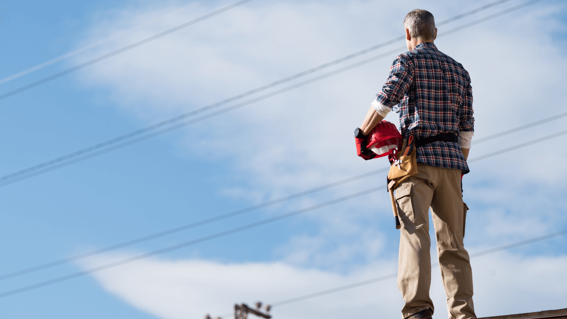 Man standing on roof holding hard hat looking at overhead electricity wires