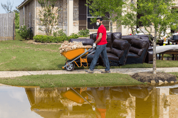 Man with wheelbarrow in face mask cleaning up after a flood, damaged furniture on sidewalk