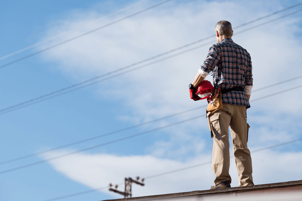 Man standing on roof holding hard hat looking at overhead electricity wires 